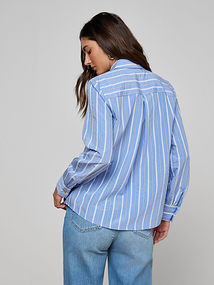 A woman with long brown hair wears the Ellery Poplin Shirt—a relaxed fit blue and white striped button-down—paired with light blue jeans, standing facing away and looking over her shoulder against a plain light gray background.