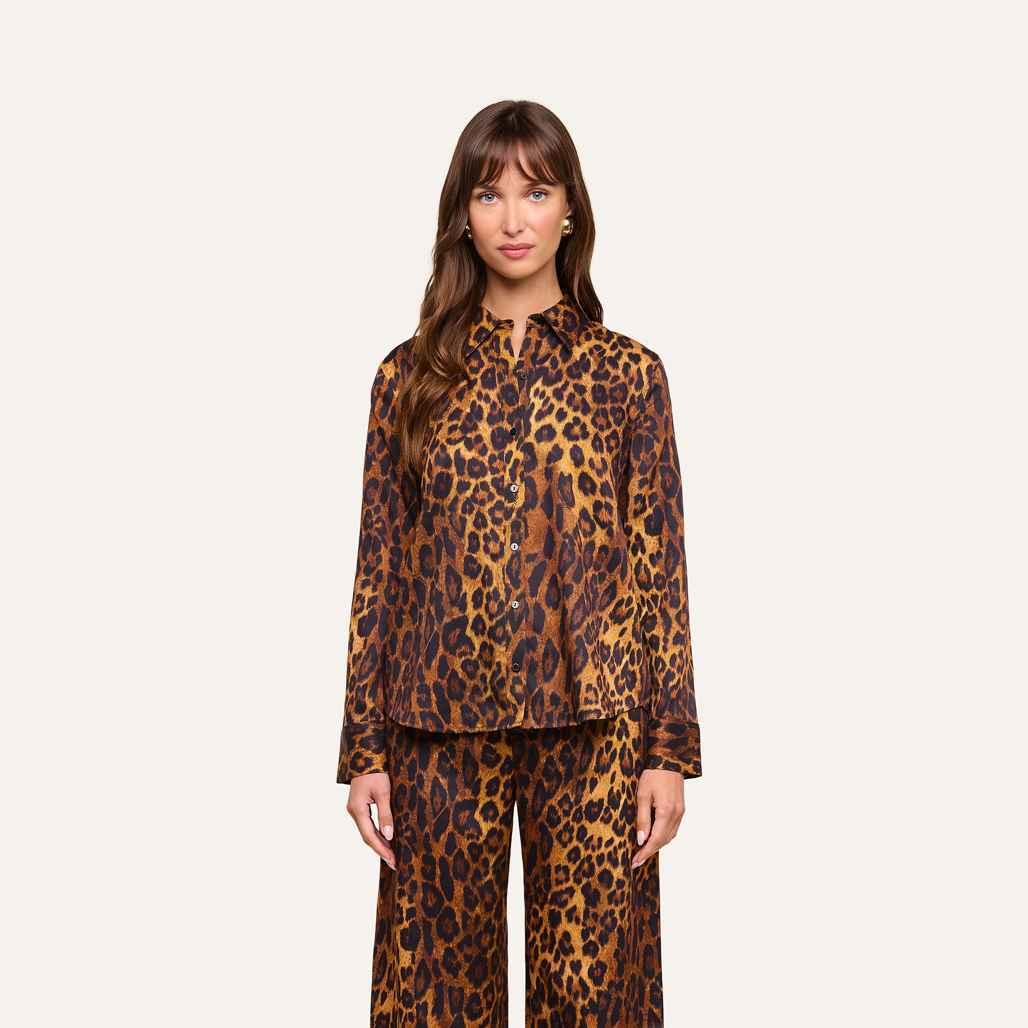 A woman stands facing forward, wearing the Ellery Blouse with long sleeves and a relaxed fit, paired with matching wide-leg pants. Both pieces feature a bold brown and black leopard print pattern. The background is plain and light-colored.