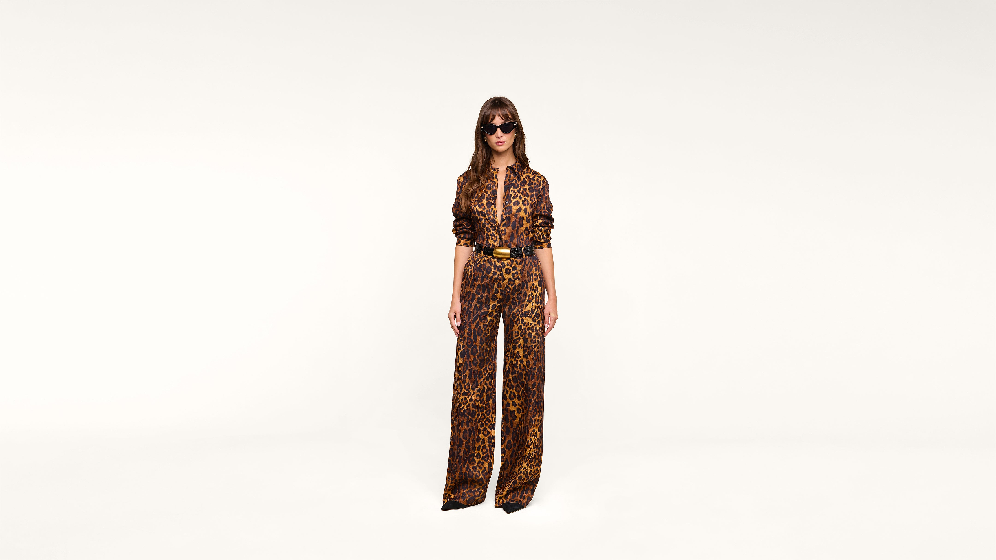 A woman wearing sunglasses and a leopard-print jumpsuit stands against a plain white background.