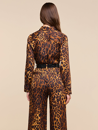 A woman with long brown hair, seen from behind, wears the Ellery Blouse—a relaxed-fit, long-sleeve leopard print top—paired with wide-leg pants and a black belt, standing against a plain beige background.