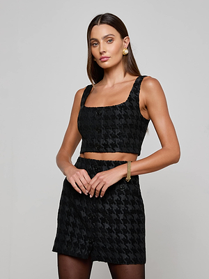 A woman with long brown hair wears the L'AGENCE Maylin Houndstooth Top and matching skirt, paired with gold earrings and a gold bracelet, as she poses against a plain gray background.