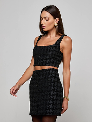 A woman models the L’AGENCE Maylin Houndstooth Top, a sleeveless black bralette crop, paired with a matching high-waisted skirt. She stands against a plain light gray background, looking down with one arm slightly extended.