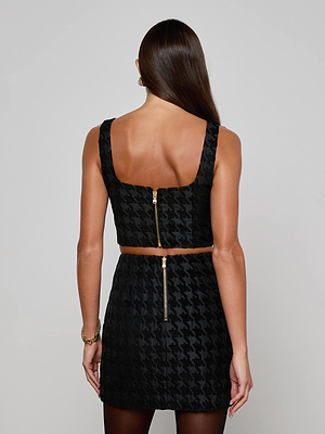 A woman with long brown hair faces away, wearing the L'AGENCE Maylin Houndstooth Top—a black sleeveless crop top with princess seams—paired with a matching high-waisted houndstooth skirt featuring gold back zippers.