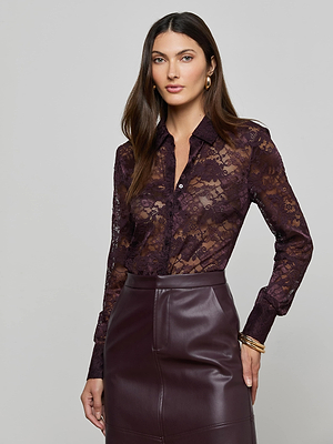 A woman wearing the L'AGENCE Maia Floral Lace Blouse in dark purple and a high-waisted burgundy leather skirt poses against a light gray background. She has long brown hair, neutral makeup, and gold hoop earrings.