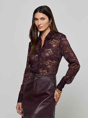 A woman wears the Maia Floral Lace Blouse by L'AGENCE in deep purple, tucked into a high-waisted dark purple leather skirt. She accessorizes with gold earrings and bracelets, her long brown hair styled against a light gray background.