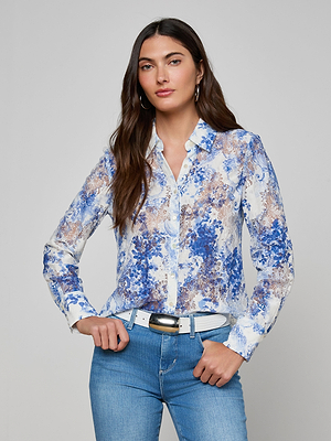 A woman with long brown hair wears the Maia Lace Blouse, paired with light blue jeans, a white belt, and hoop earrings. She poses against a plain light gray background with her hands on her hips.