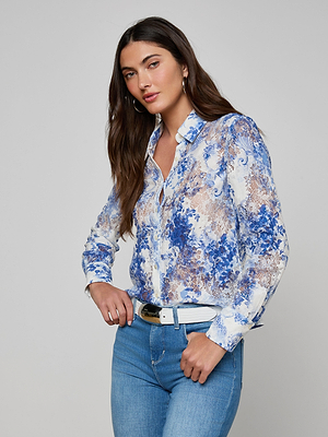 A woman with long brown hair wears the Maia Lace Blouse in navy with a floral design, paired with light blue jeans, a white belt, and hoop earrings, standing against a plain gray background.