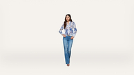 Woman standing against a plain white background, wearing a blue and white floral blouse, blue jeans, and beige heels, with her hands resting on her hips and looking forward.