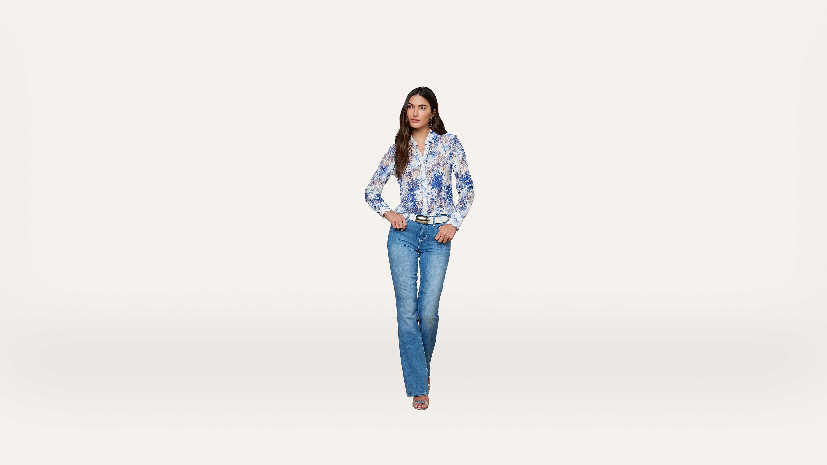 Woman standing against a plain white background, wearing a blue and white floral blouse, blue jeans, and beige heels, with her hands resting on her hips and looking forward.