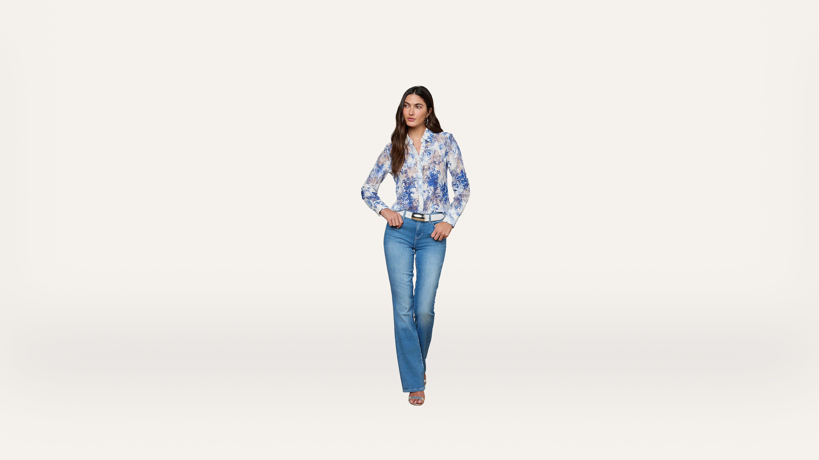Woman standing against a plain white background, wearing a blue and white floral blouse, blue jeans, and beige heels, with her hands resting on her hips and looking forward.