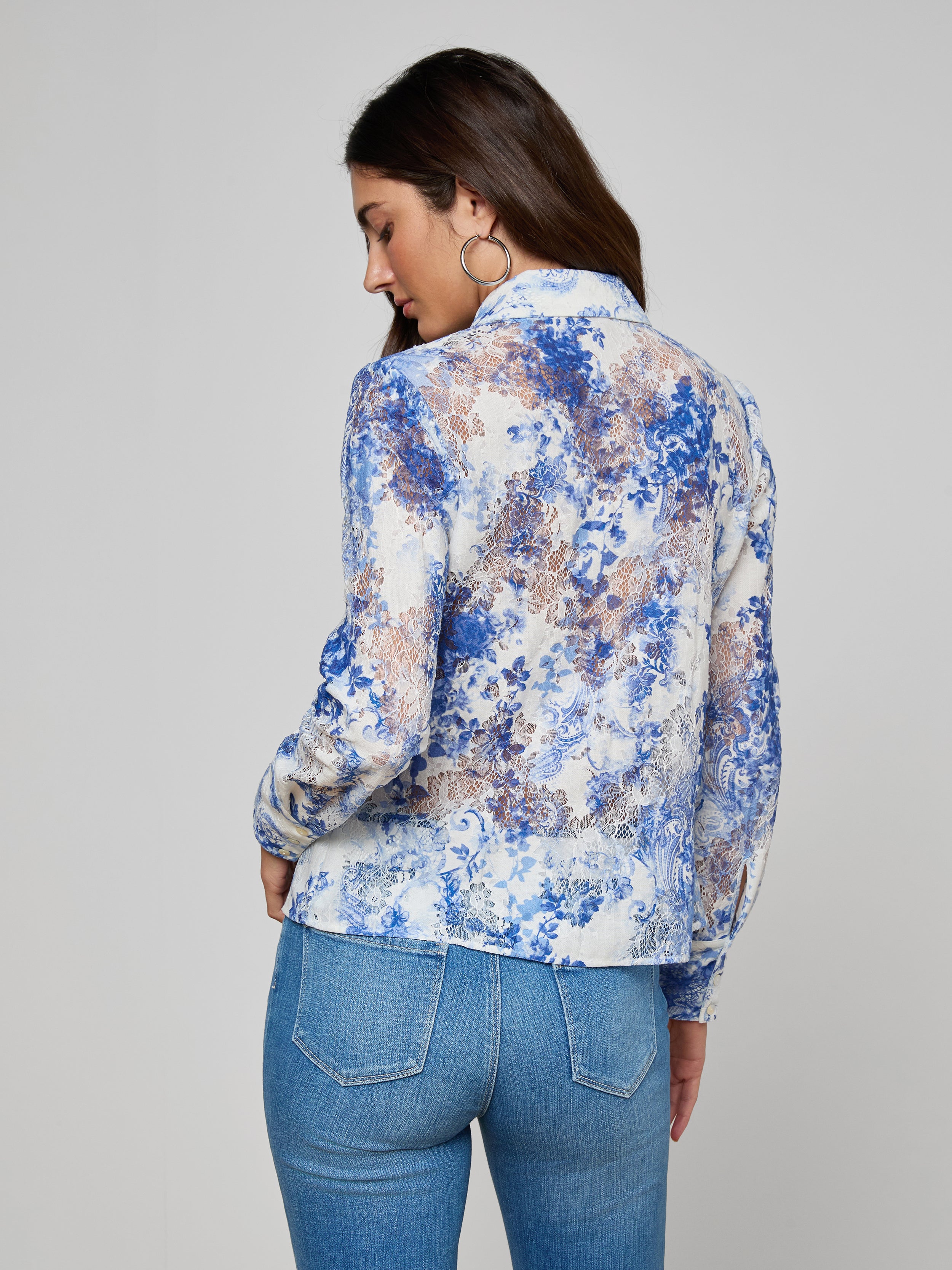 A woman with long brown hair, large hoop earrings, and blue jeans models the Maia Lace Blouse, standing against a plain light gray background while looking over her shoulder.