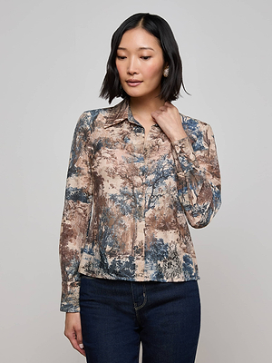 A woman with short dark hair wears the Maia Printed Lace Blouse by L'AGENCE, featuring a long-sleeved Western toile print in blue and brown, standing against a plain light background while looking down and touching her neck.