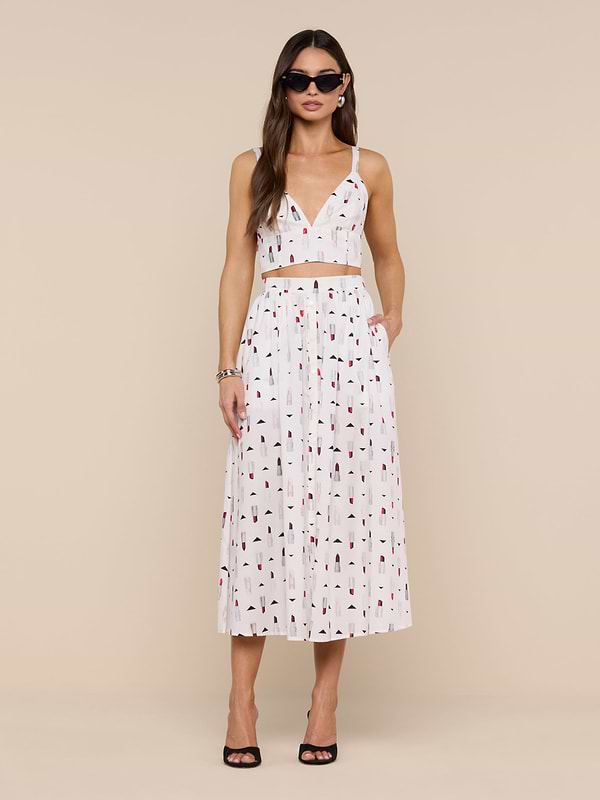 A woman wearing sunglasses and a white two-piece outfit featuring the Kaori Bralette Top with a lipstick print and a matching midi skirt, standing against a plain white background. She completes the look with black heeled sandals and gold jewelry.