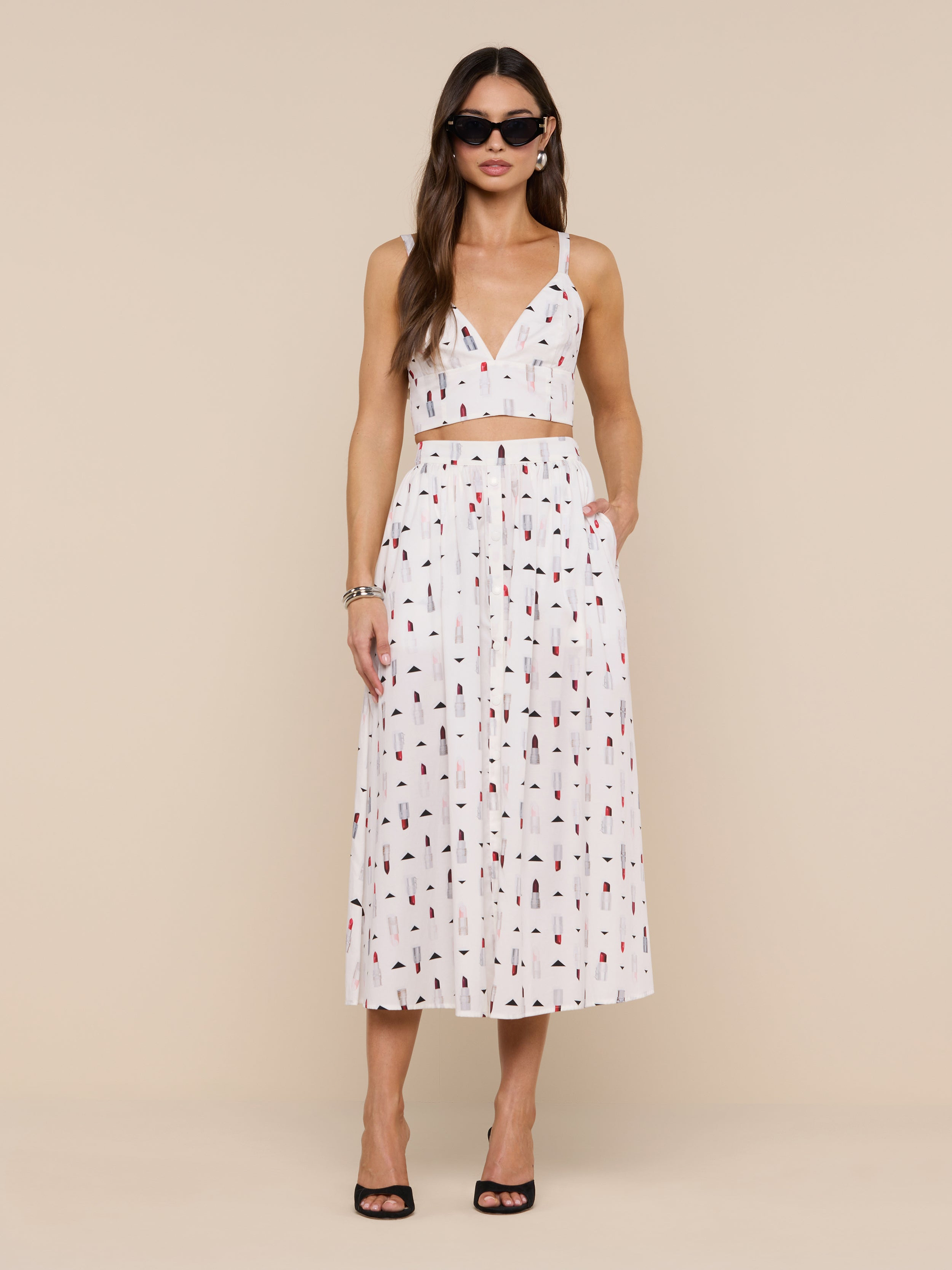 A woman wearing sunglasses and a white two-piece outfit featuring the Kaori Bralette Top with a lipstick print and a matching midi skirt, standing against a plain white background. She completes the look with black heeled sandals and gold jewelry.
