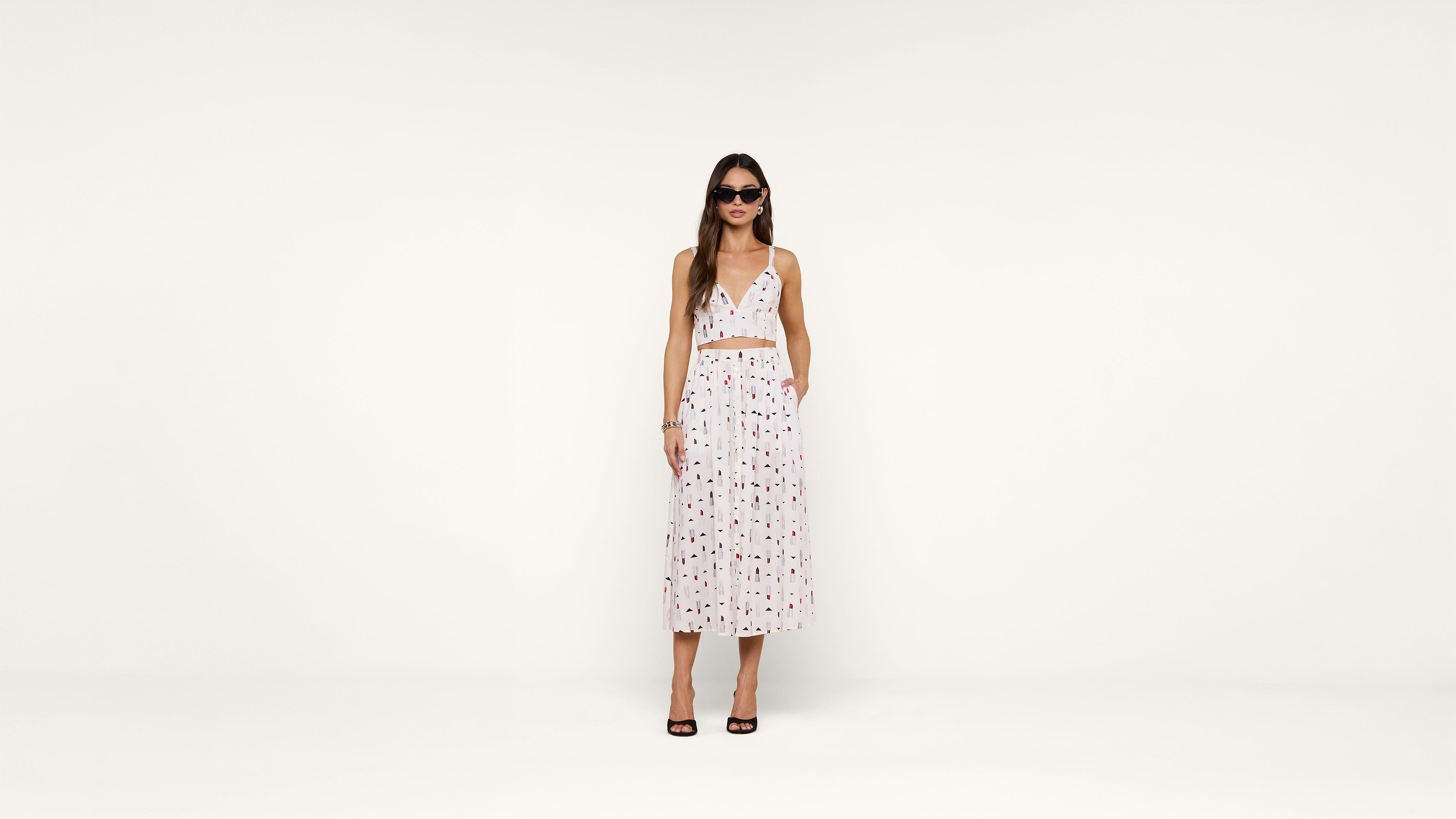 A woman in sunglasses wears a white sleeveless crop top and matching high-waisted, ankle-length skirt with a red and black pattern, standing against a plain white background.