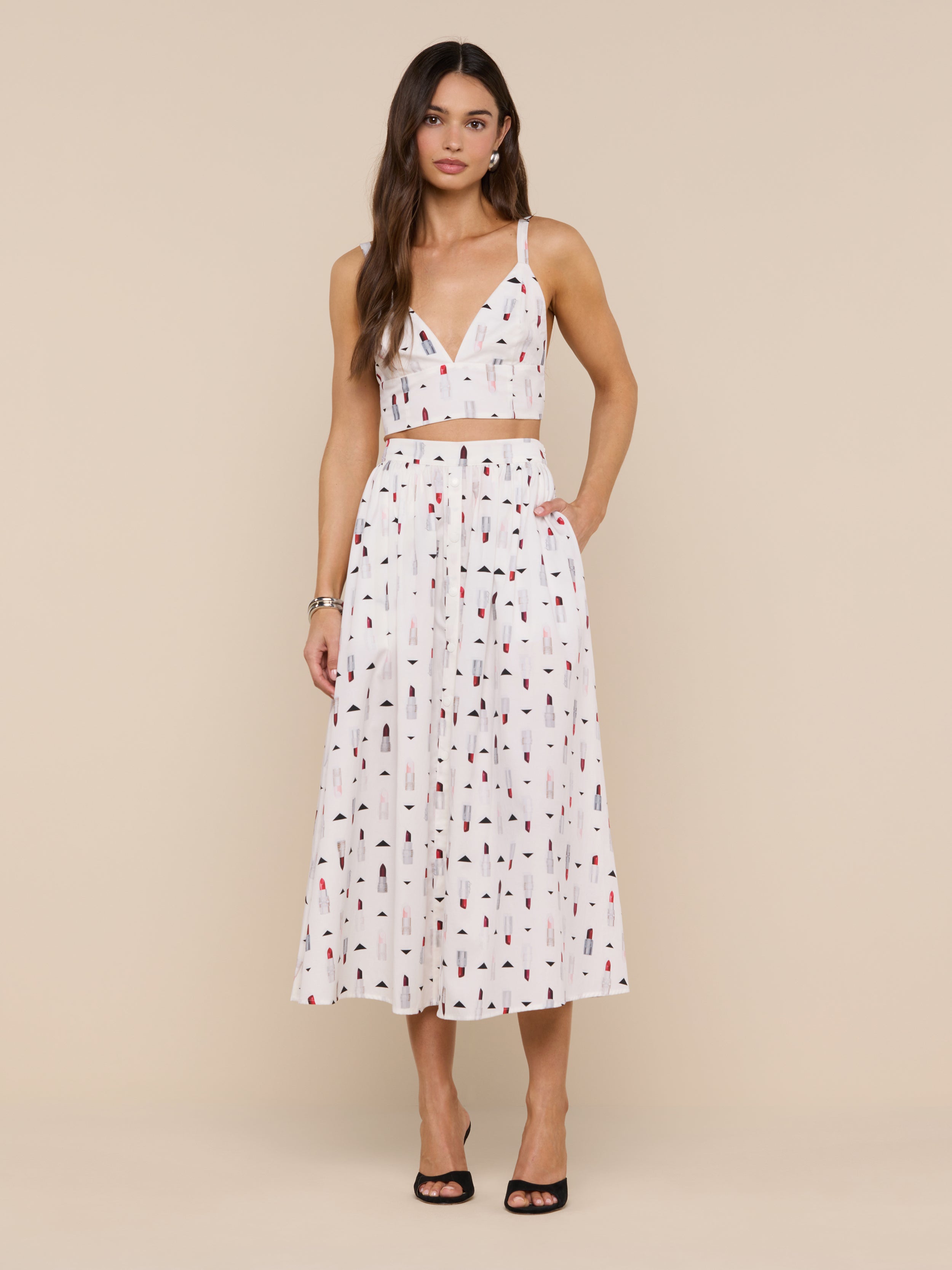 Woman wearing the Kaori Bralette Top styled with a white pleated midi skirt featuring small multicolored lipstick prints and a wide waist band, standing against a plain white background and pairing the look with black open-toe heels.