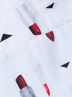 The Kaori Bralette Top features white poplin fabric with a repeating lipstick print of red tubes and small black triangles, shown folded through the center of the image.