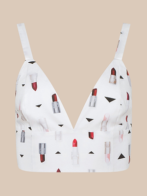 The Kaori Bralette Top features thin straps, a deep V neckline, and a playful lipstick print with small black triangles on soft poplin fabric, shown against a plain beige background.