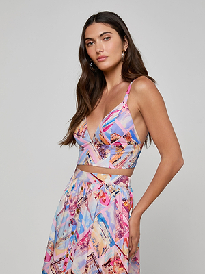 A woman with long brown hair wears the Kaori Bralette Top and a matching high-waisted skirt in a colorful geometric print. She stands against a plain light gray background, looking confidently at the camera with one hand on her hip.