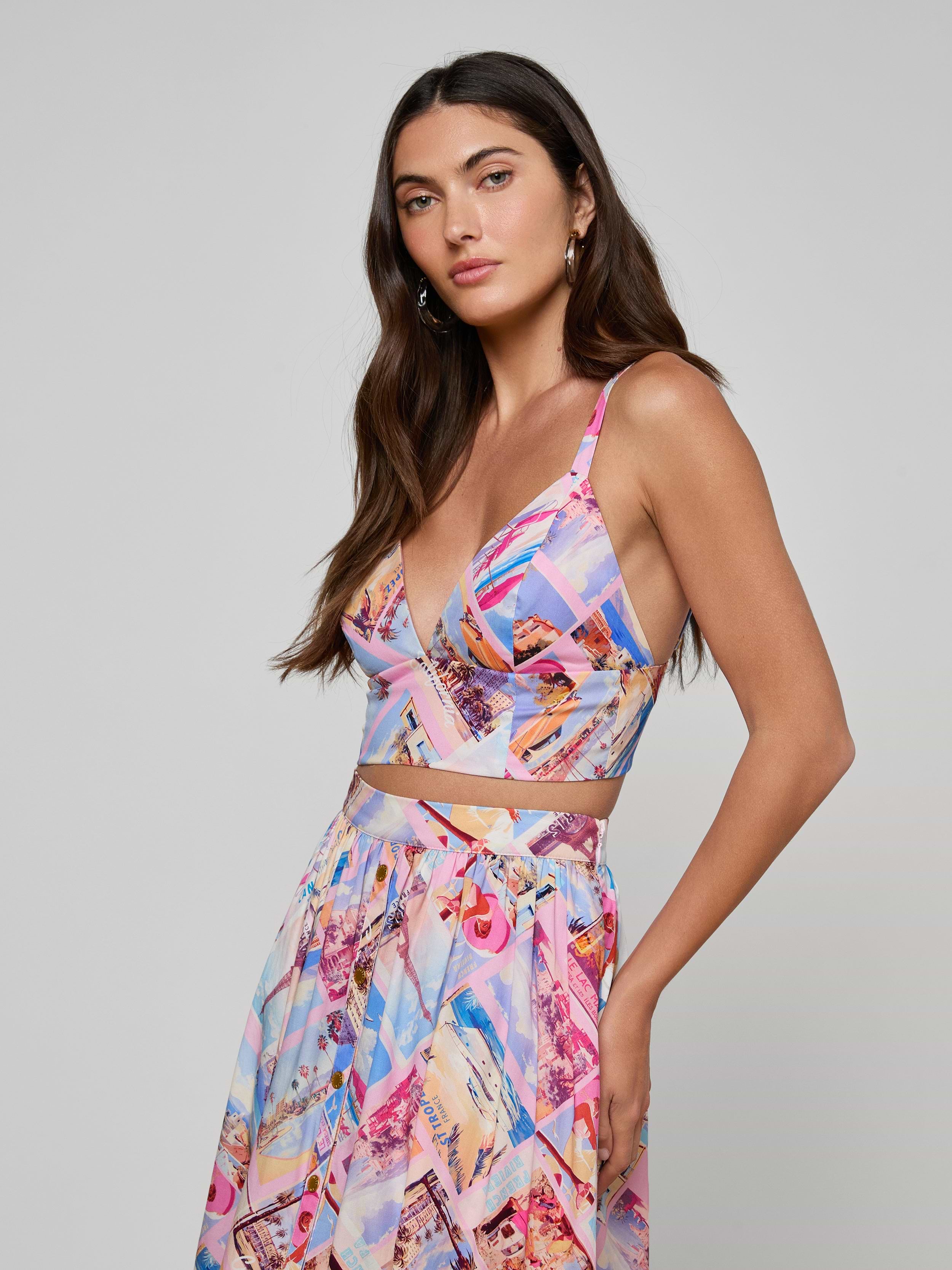 A woman with long, loosely styled dark hair stands confidently against a plain light background, wearing the Kaori Bralette Top in a colorful postcard print, paired with a matching skirt crafted from cotton-blend poplin.