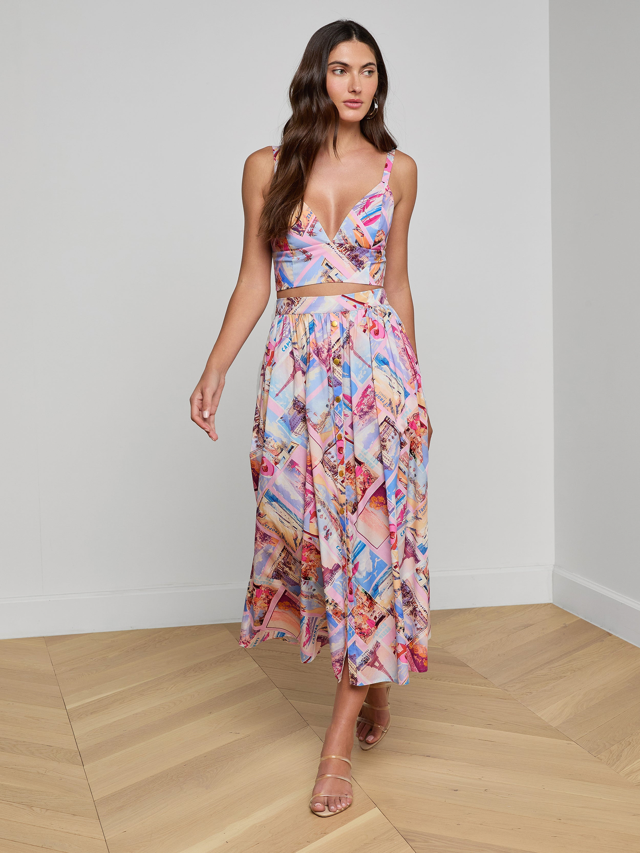 A woman styled in the Kaori Bralette Top and a vibrant postcard print midi skirt, both crafted from cotton-blend poplin, paired with nude heels as she walks against a plain white background.