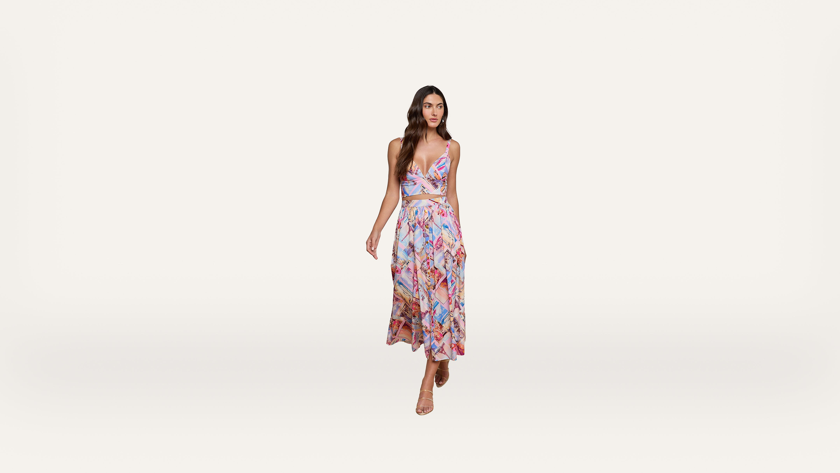 A woman in a colorful, patterned, sleeveless dress with a cutout at the waist stands against a plain white background, wearing nude heels.