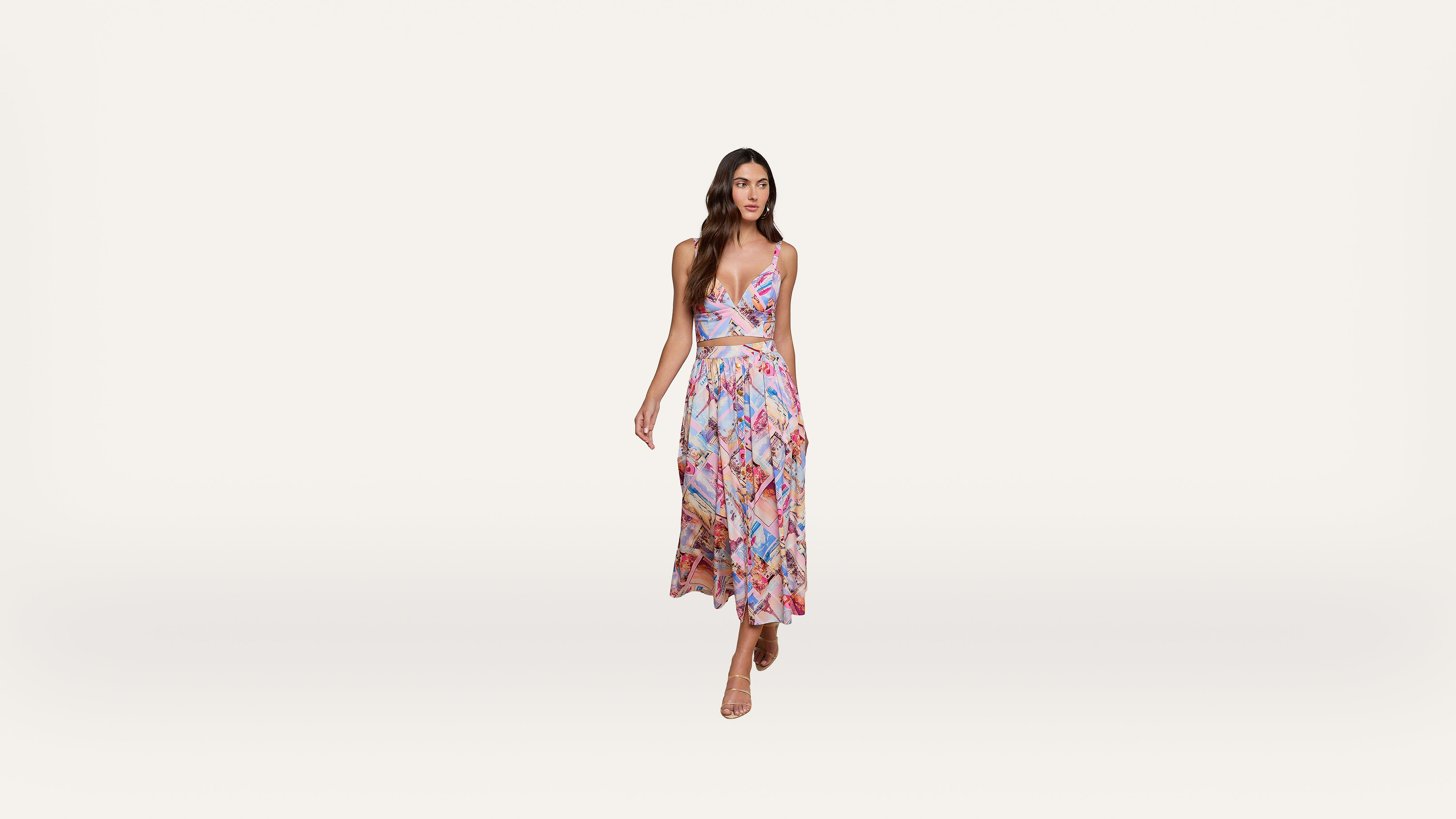 A woman in a colorful, patterned, sleeveless dress with a cutout at the waist stands against a plain white background, wearing nude heels.