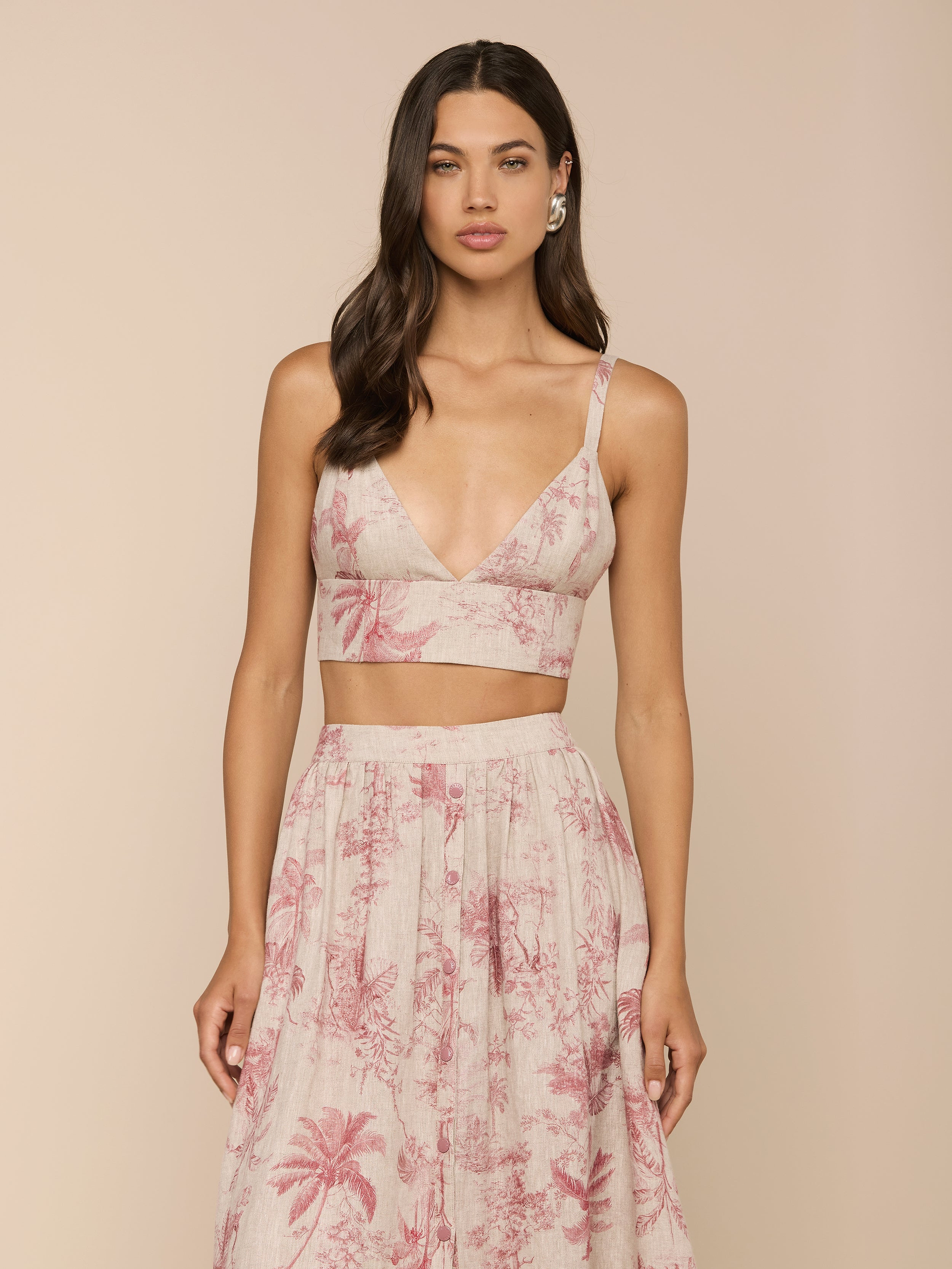 Wearing the Kaori Linen Bralette Top in light pink, a woman stands against a plain light background. She pairs the thin-strapped bralette with a matching high-waisted skirt featuring a palm tree pattern.