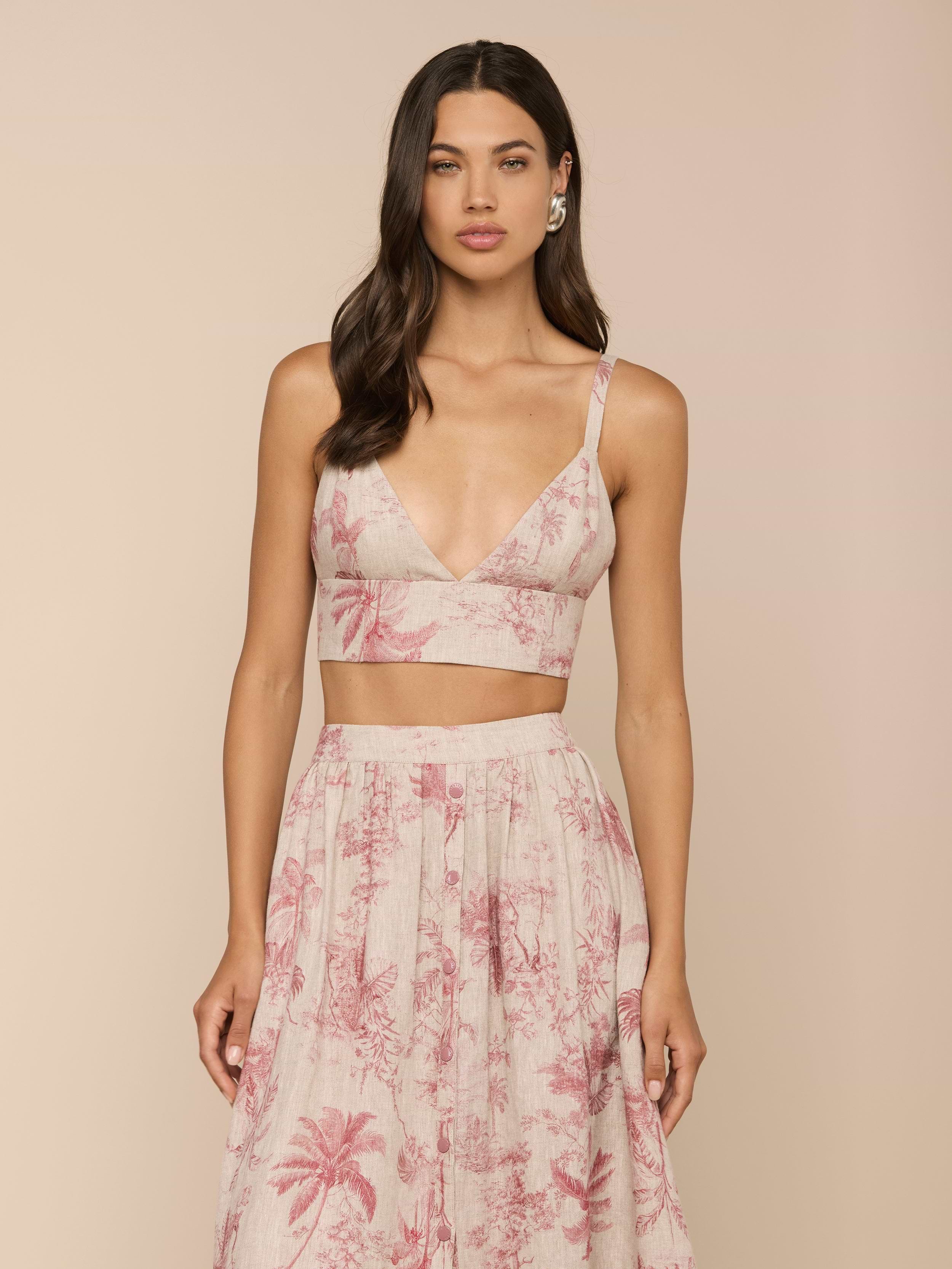 Wearing the Kaori Linen Bralette Top in light pink, a woman stands against a plain light background. She pairs the thin-strapped bralette with a matching high-waisted skirt featuring a palm tree pattern.
