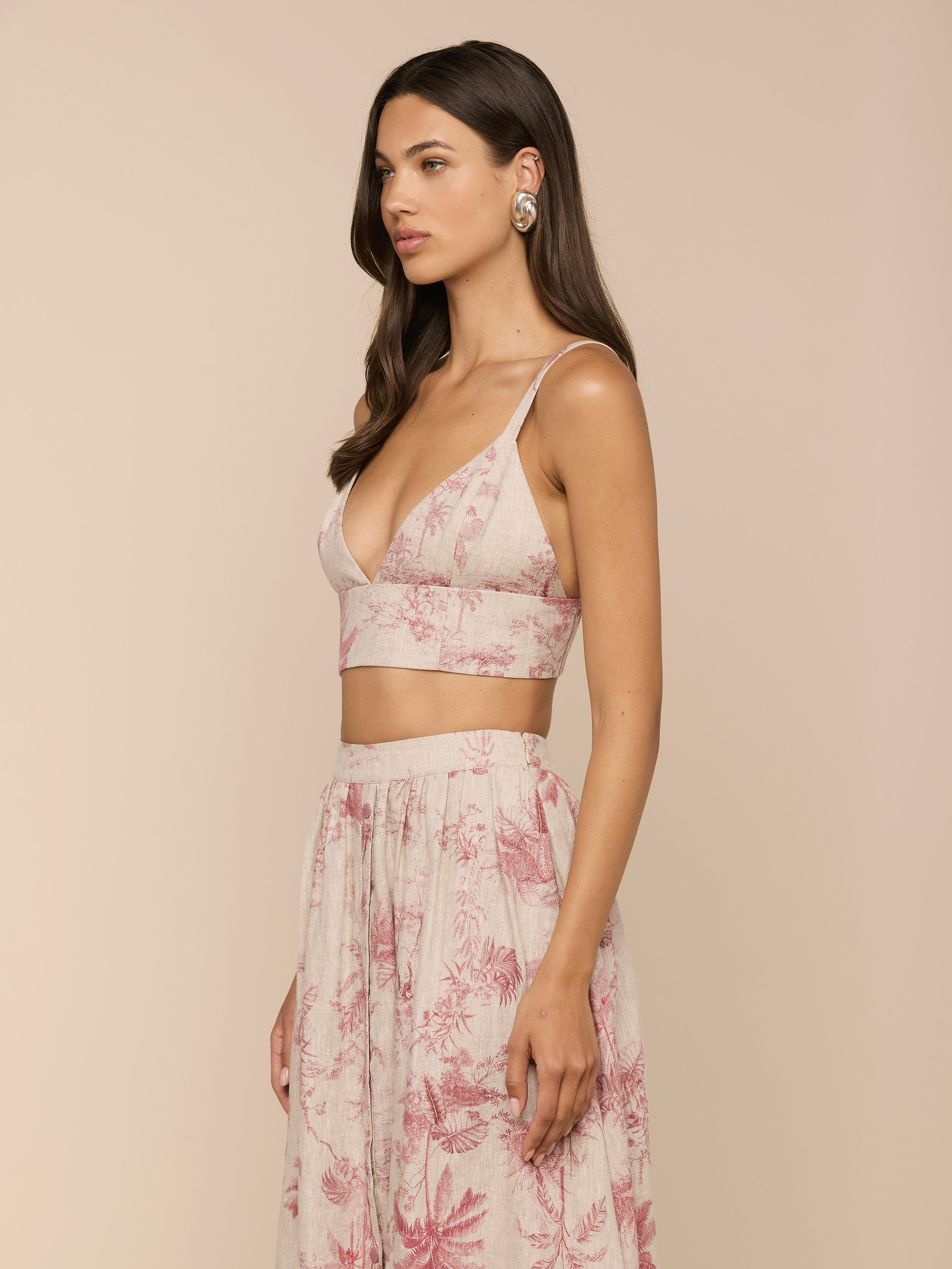 A woman with long brown hair wears the Kaori Linen Bralette Top in pink and cream floral, paired with a high-waisted skirt. She is standing sideways against a plain white background.