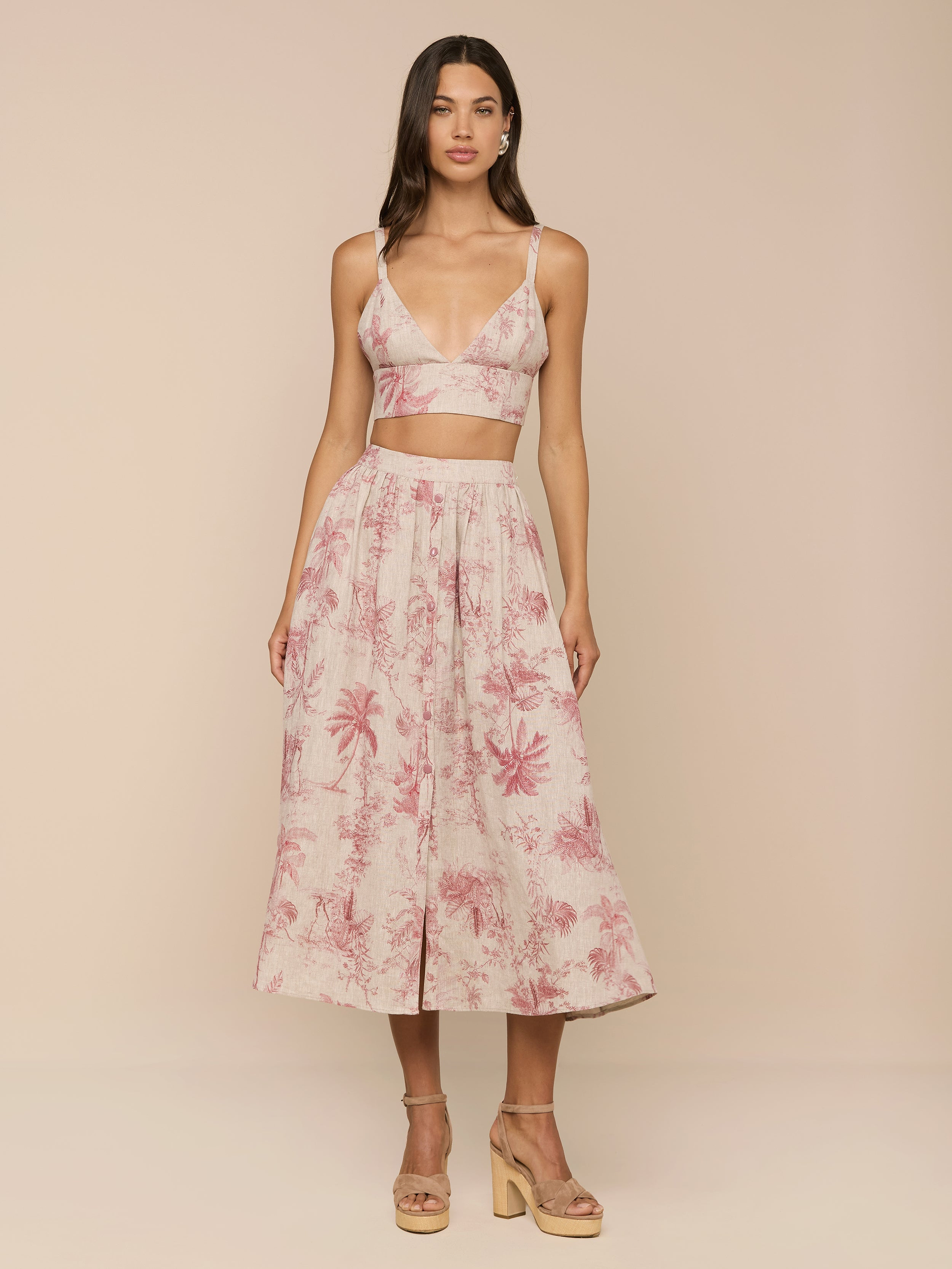 A woman stands in a studio wearing the light pink Kaori Linen Bralette Top in a jungle toile print, paired with a matching midi skirt and tan platform sandals.