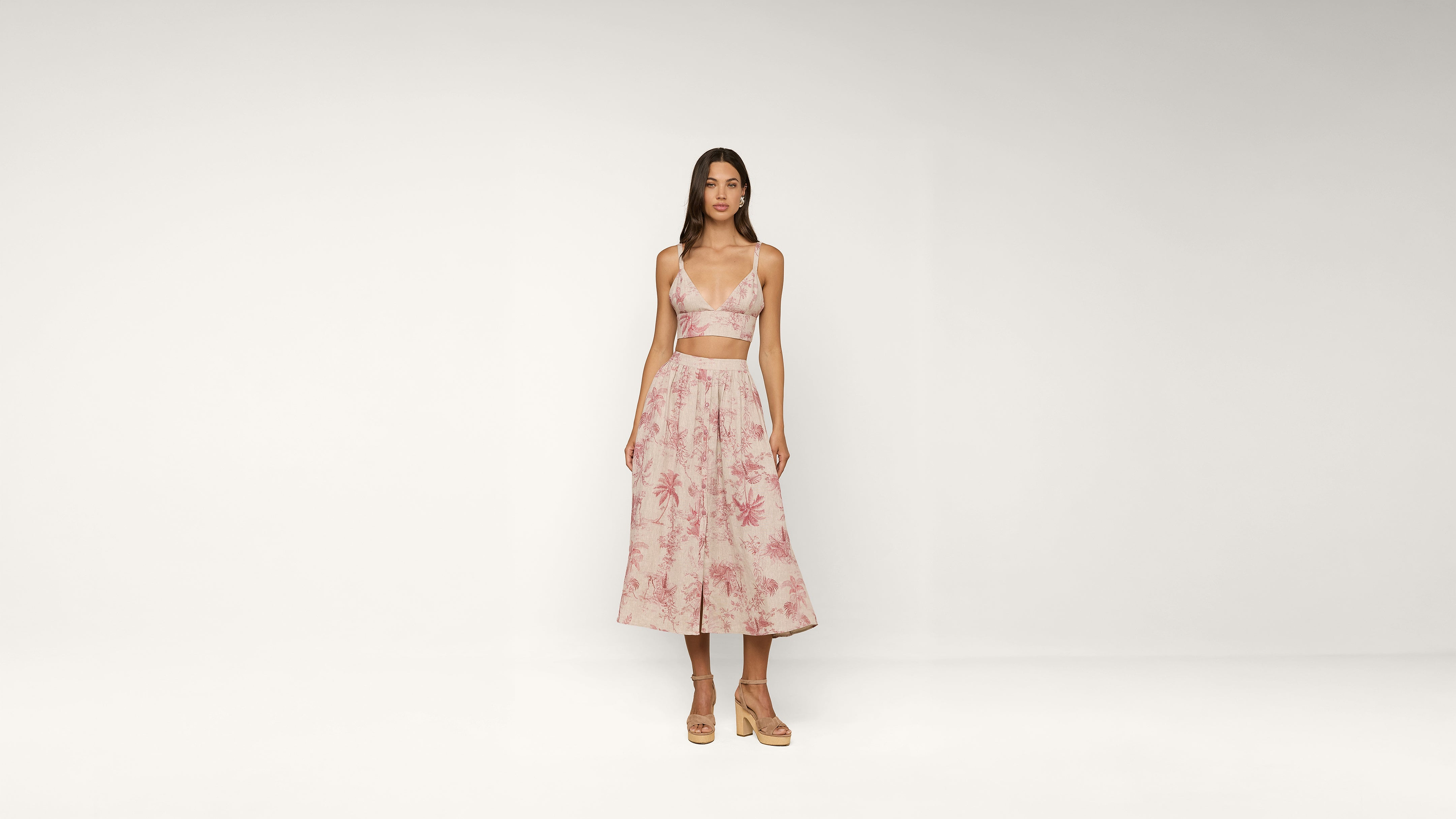 A woman stands against a plain white background, wearing a pink floral crop top and matching midi skirt with sandals. She faces forward with her arms at her sides.