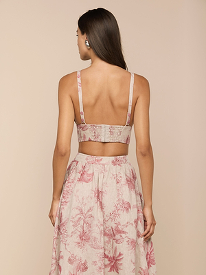 A woman with long brown hair stands facing away, wearing the Kaori Linen Bralette Top and a high-waisted skirt with a pink floral pattern on beige. The backdrop is plain, light beige.