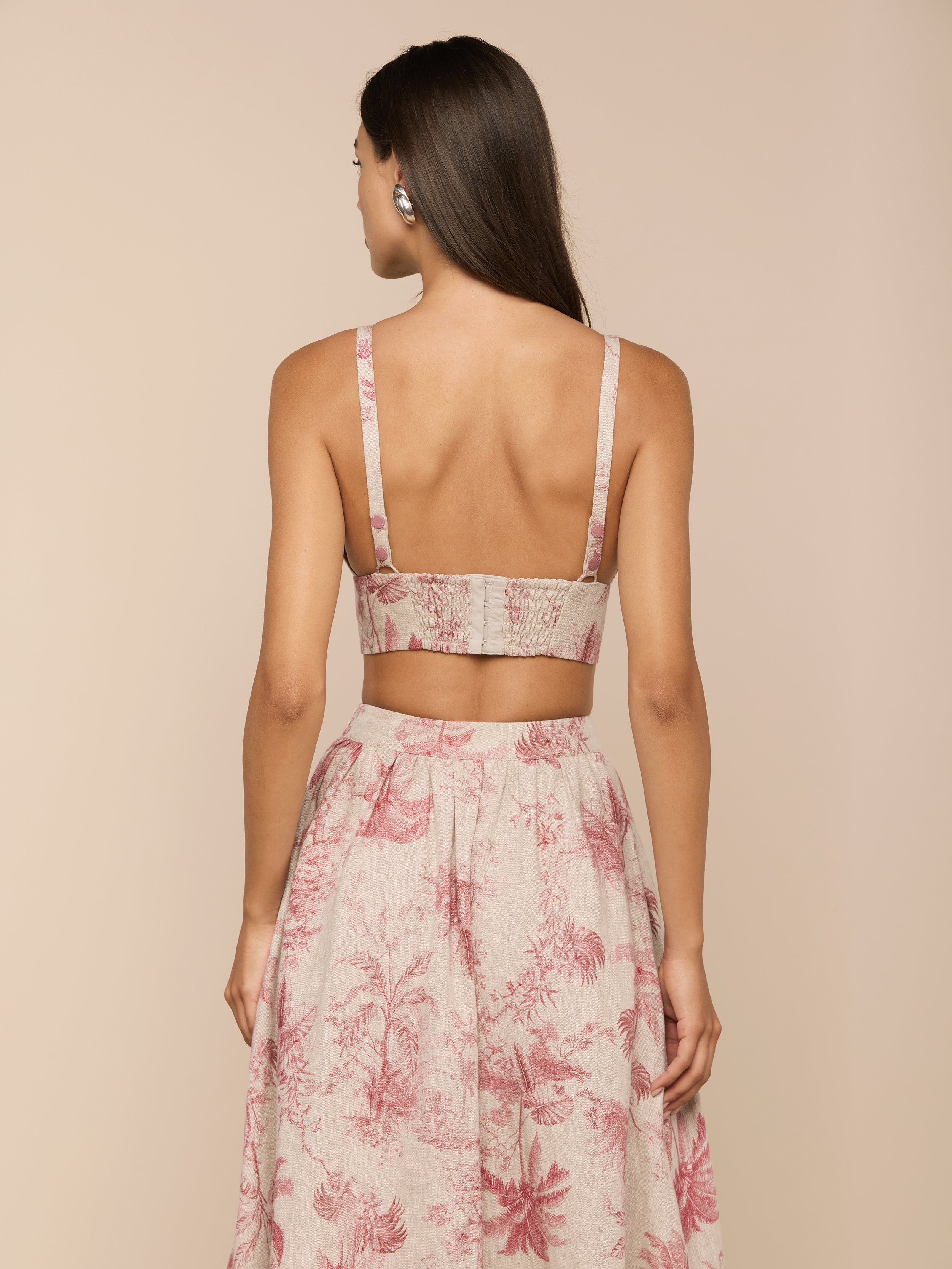 A woman with long brown hair, wearing the sleeveless Kaori Linen Bralette Top in a jungle toile print and a matching high-waisted skirt, stands facing away with her hands at her sides against a plain light background.
