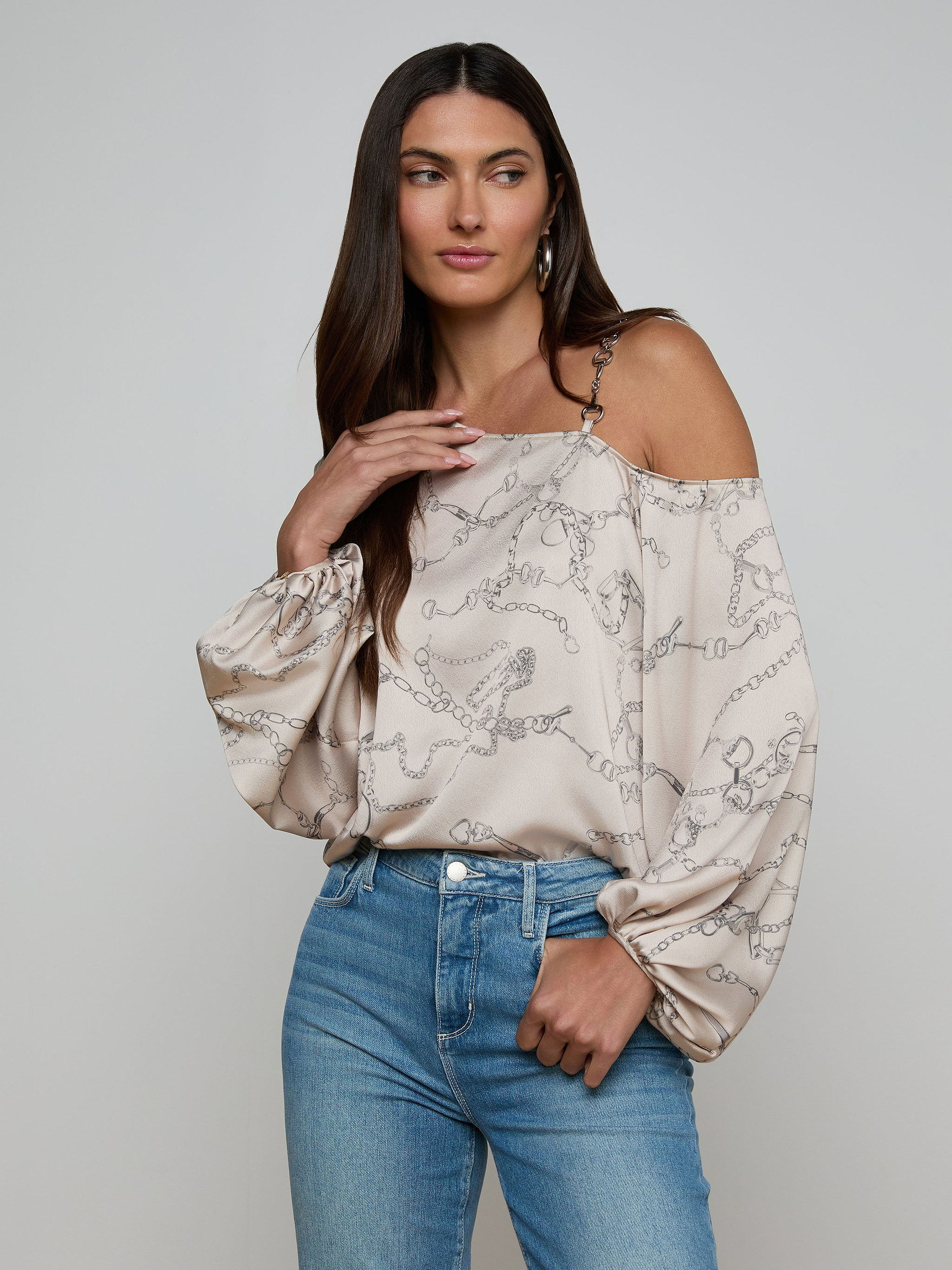A woman wearing the Asami Chain One-Shoulder Blouse in light beige satin with horse bit chain patterns and puffy sleeves, paired with light blue jeans, stands against a plain background.