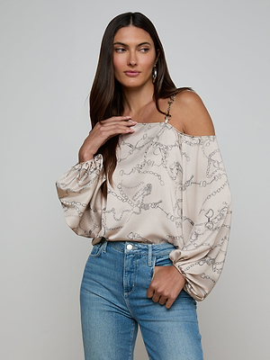 A woman with long brown hair wears the beige Asami Chain One-Shoulder Blouse featuring horse bit chain patterns and light blue jeans. She poses with a hand near her shoulder, gazing to the side against a plain light background.
