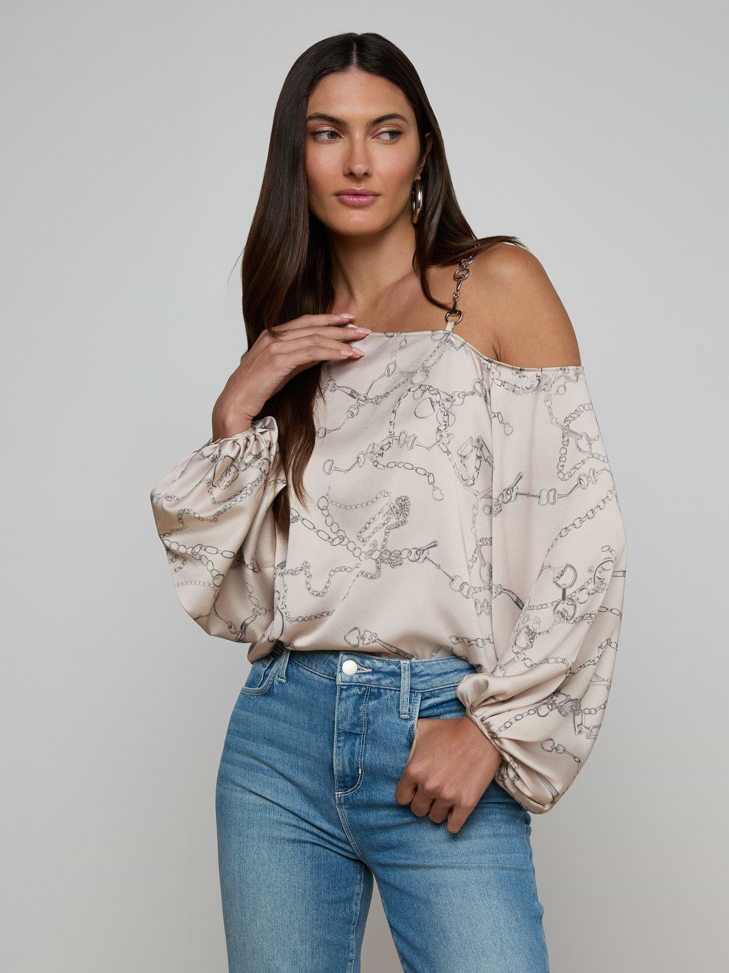 A woman wearing the Asami Chain One-Shoulder Blouse in light beige satin with horse bit chain patterns and puffy sleeves, paired with light blue jeans, stands against a plain background.