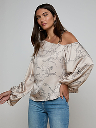 A woman poses against a plain light background wearing the Asami Chain One-Shoulder Blouse, a loose beige top featuring a horse bit chain print and long puffy sleeves, paired with blue jeans.