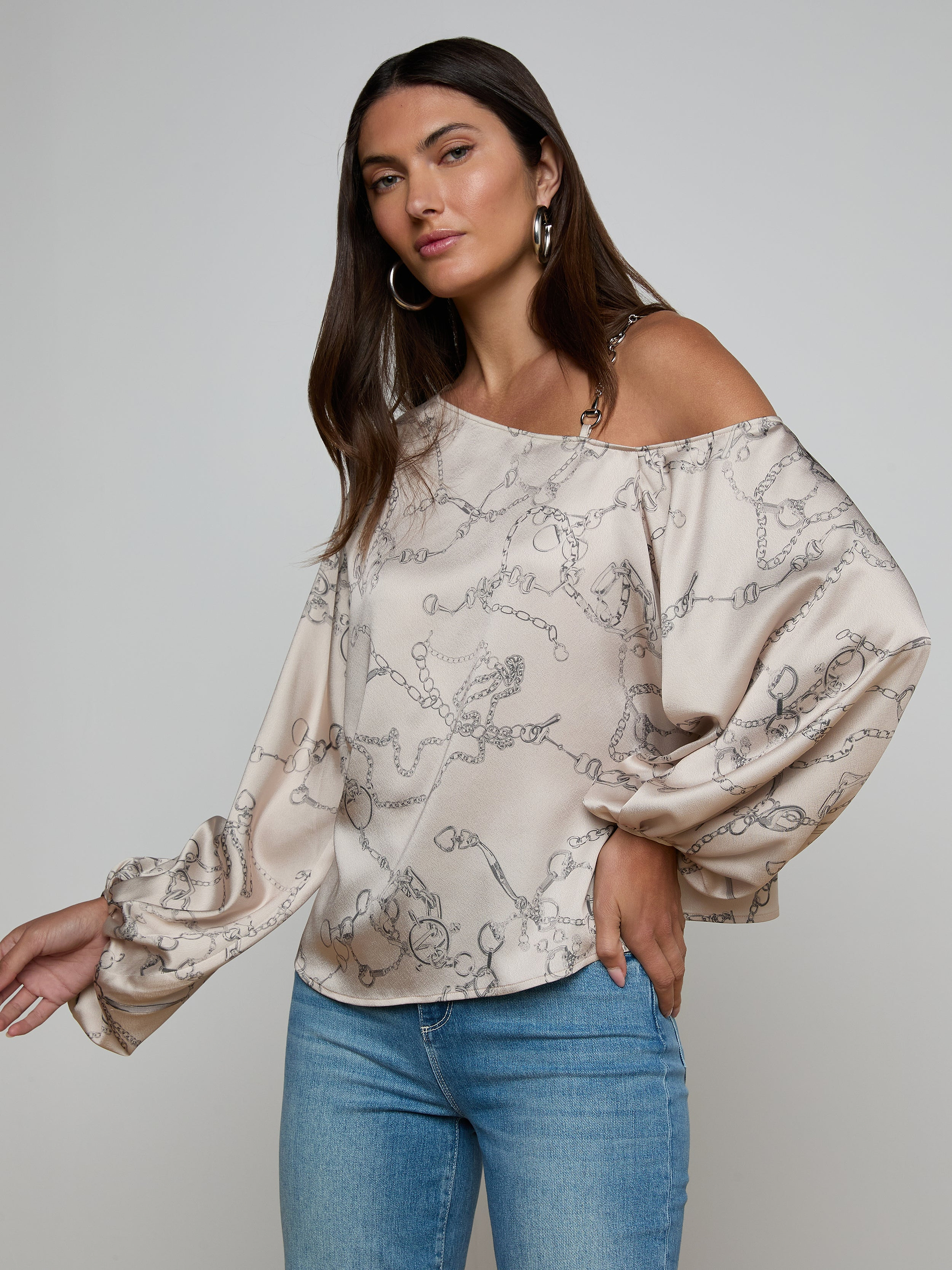 A woman poses against a plain light background wearing the Asami Chain One-Shoulder Blouse, a loose beige top featuring a horse bit chain print and long puffy sleeves, paired with blue jeans.