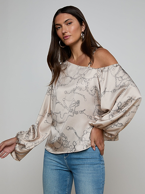 A woman with long brown hair wears the Asami Chain One-Shoulder Blouse, featuring a cream satin fabric with horse bit chain print and loose sleeves, paired with light blue jeans. She poses against a plain light background, one hand raised.