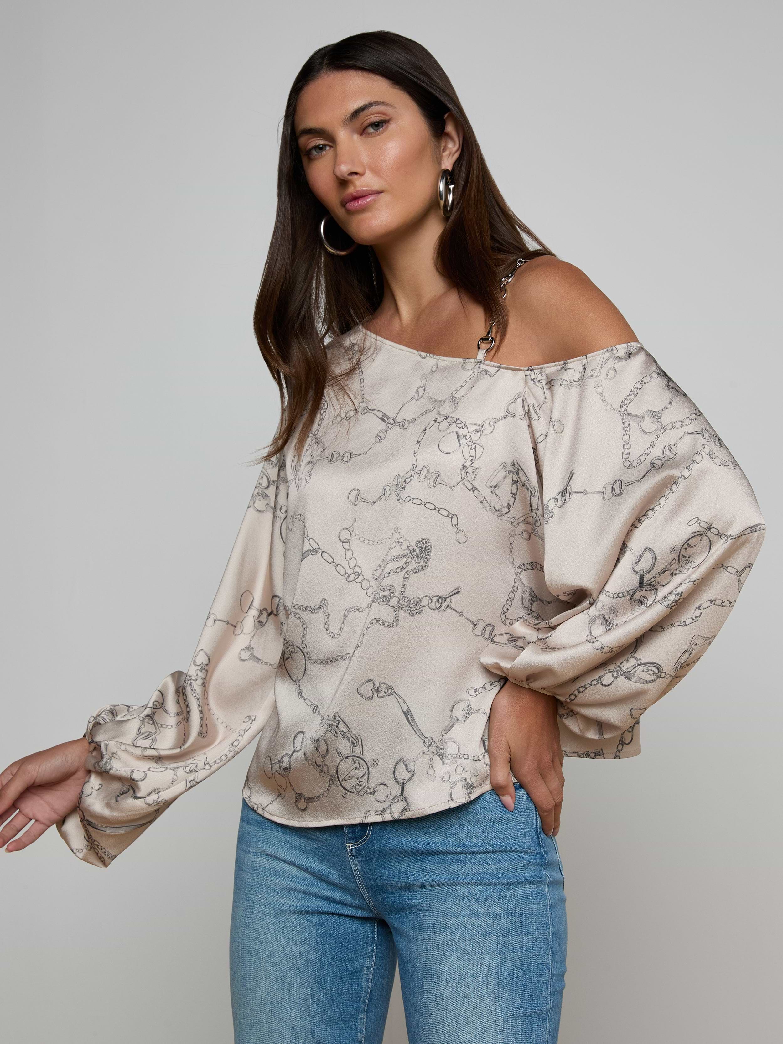 A woman poses against a plain light background wearing the Asami Chain One-Shoulder Blouse, a loose beige top featuring a horse bit chain print and long puffy sleeves, paired with blue jeans.