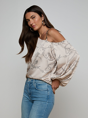 A woman with long brown hair and hoop earrings wears the Asami Chain One-Shoulder Blouse in beige with a horse bit chain print, paired with light blue high-waisted jeans, posing against a plain light background.
