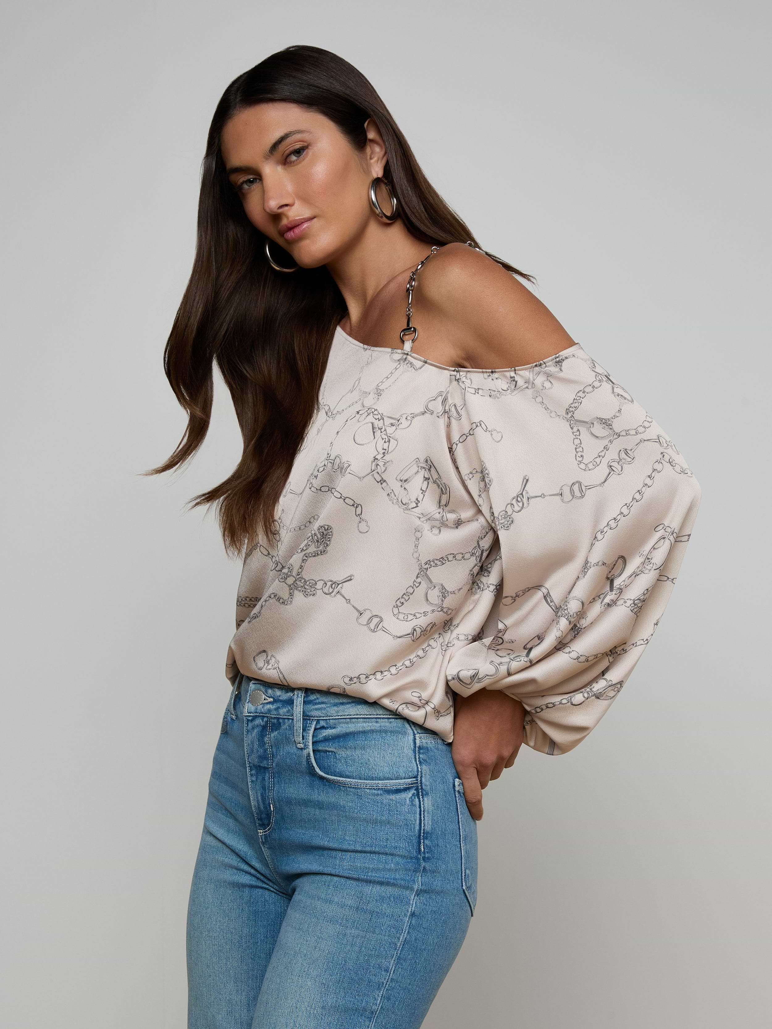 Woman with long dark hair wears the Asami Chain One-Shoulder Blouse and blue jeans, standing against a plain background with one hand in her pocket, looking slightly to the side.