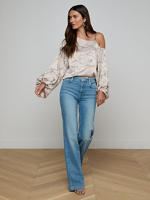 A woman with long brown hair stands on a wooden floor, looking to the side with one hand in her pocket. She wears light blue high-waisted jeans, heels, and the Asami Chain One-Shoulder Blouse with long sleeves.