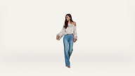 Woman wearing a floral off-shoulder blouse and high-waisted blue jeans stands against a plain white background, looking to her left with a relaxed expression.