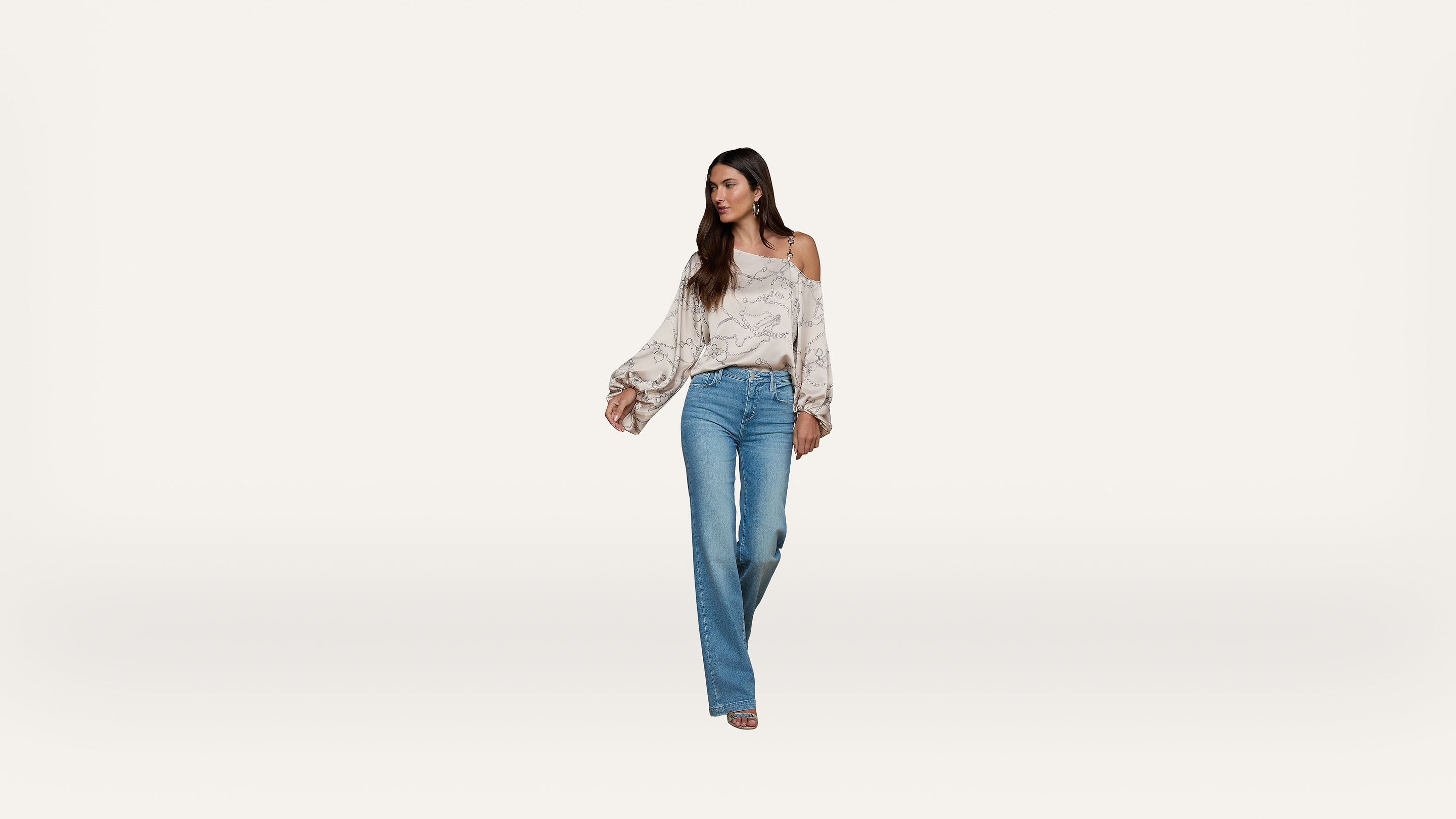 Woman wearing a floral off-shoulder blouse and high-waisted blue jeans stands against a plain white background, looking to her left with a relaxed expression.
