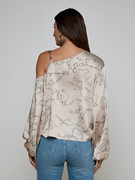 A woman with long brown hair is seen from the back against a plain background, wearing the Asami Chain One-Shoulder Blouse—a light-colored satin top featuring a horse bit chain pattern—paired with blue jeans.