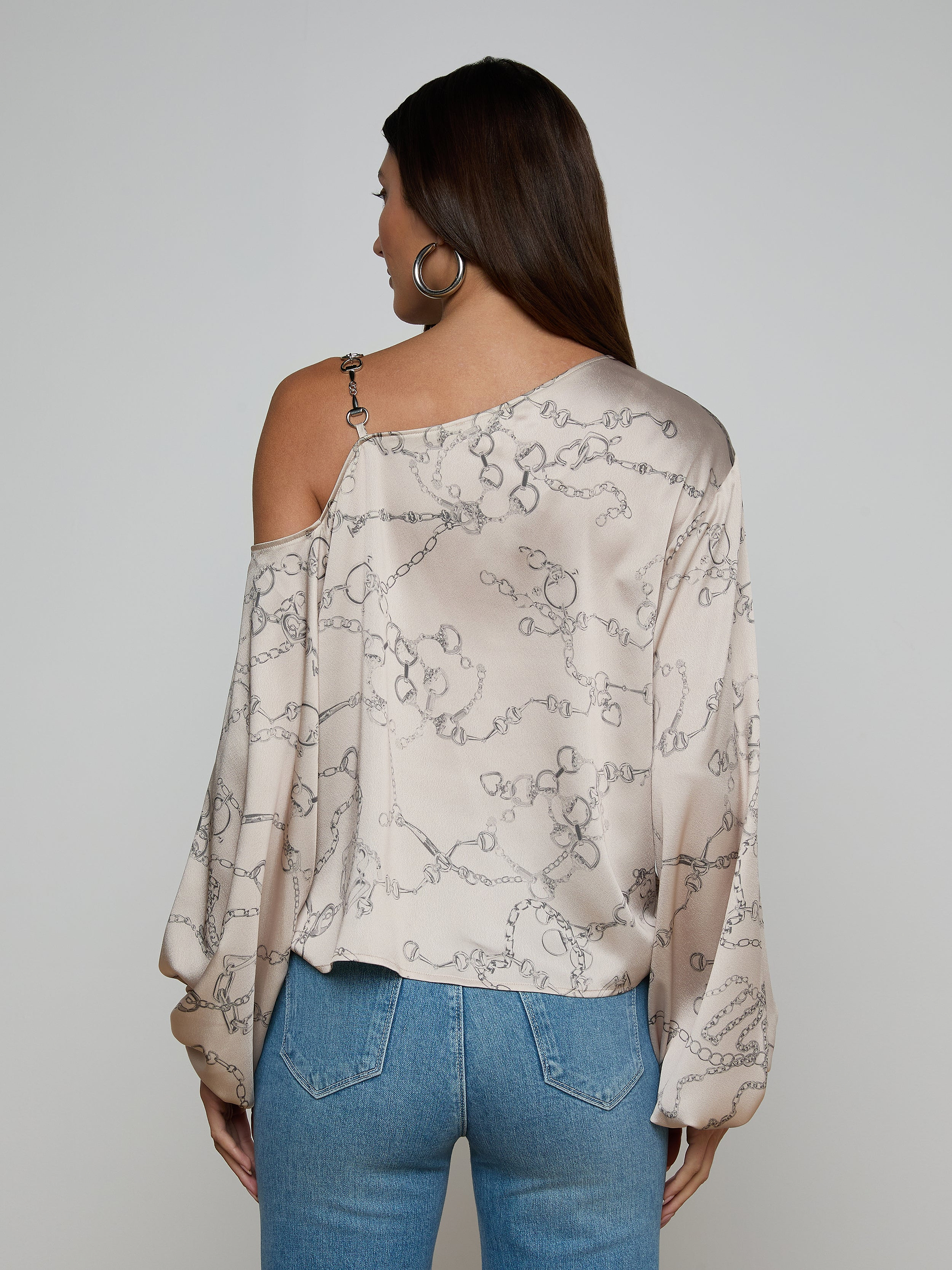 A woman with long brown hair is seen from the back against a plain background, wearing the Asami Chain One-Shoulder Blouse—a light-colored satin top featuring a horse bit chain pattern—paired with blue jeans.