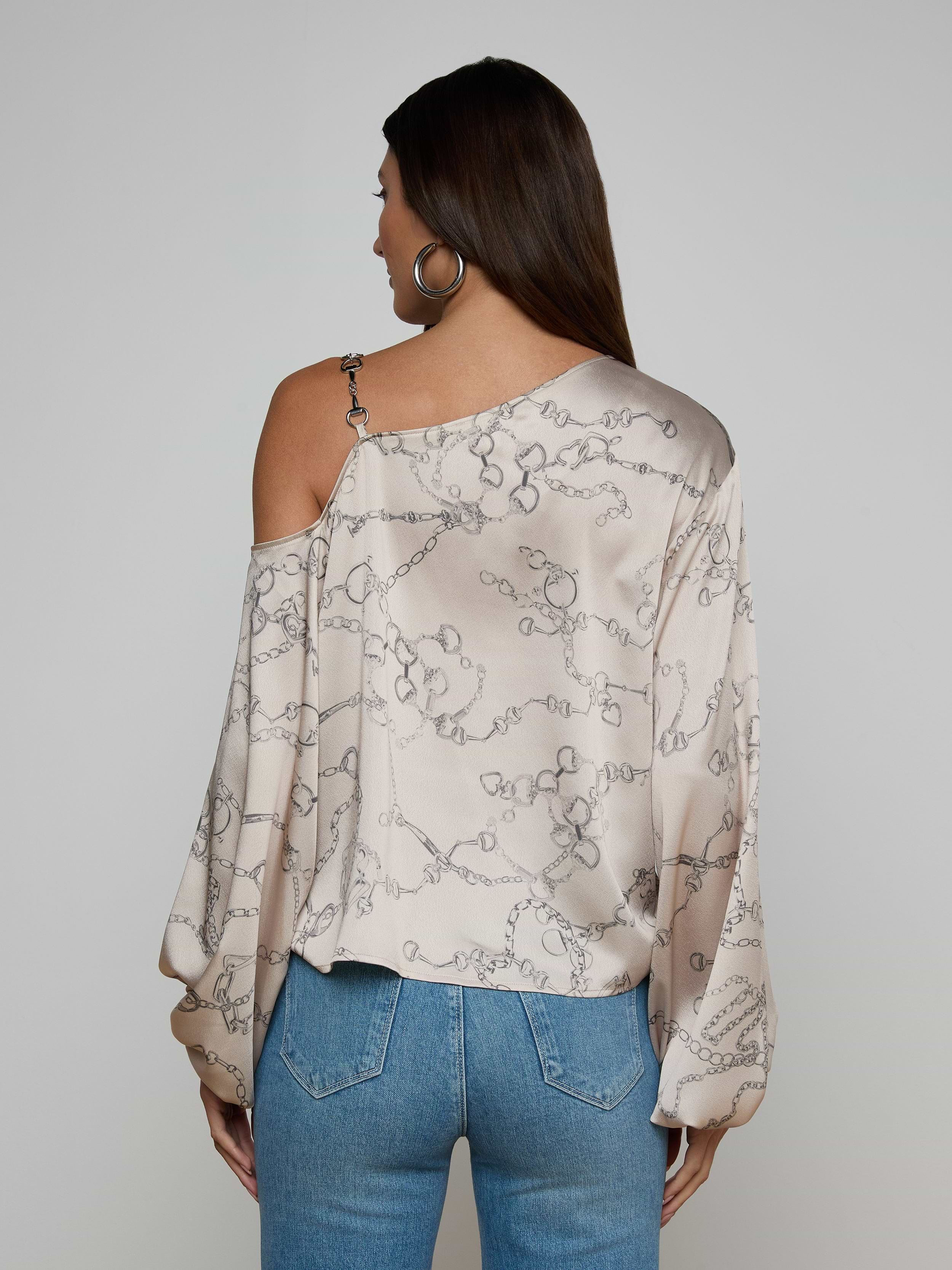 A woman with long brown hair is seen from the back against a plain background, wearing the Asami Chain One-Shoulder Blouse—a light-colored satin top featuring a horse bit chain pattern—paired with blue jeans.
