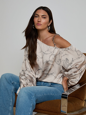 A woman with long brown hair sits on a brown chair, wearing the Asami Chain One-Shoulder Blouse—an off-shoulder cream satin top with a horse bit chain pattern—paired with blue jeans and hoop earrings, looking relaxed and confident.